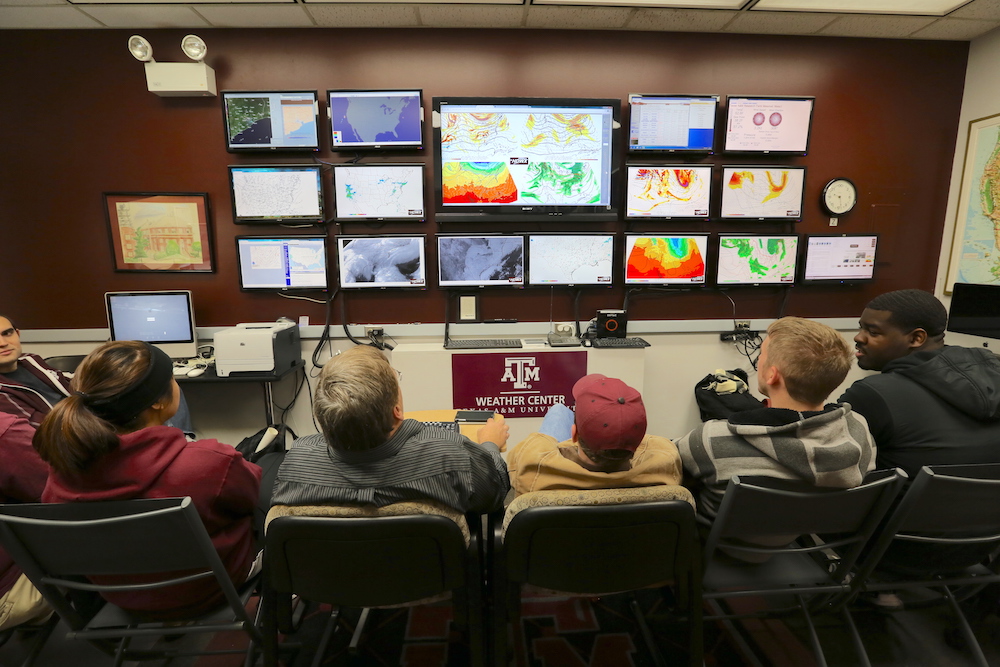 Six students seated next to each other and looking at several monitors on a wall that display weather patterns.