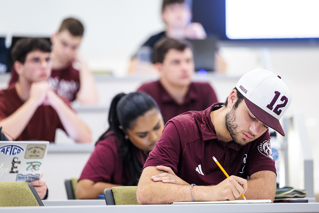 Male student wearing maroon and white hat takes notes in classroom