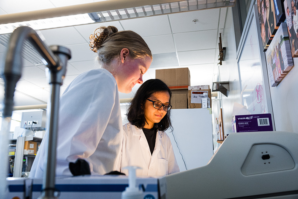 Two female students in lab coats looking into lit screen in lab setting