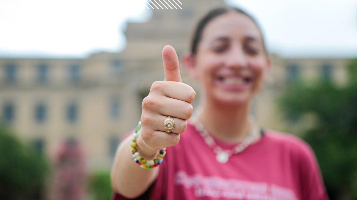 A student giving a thumbs up with the focus on their hand. Their face is blurred behind it, and they are wearing a gold Aggie Ring.