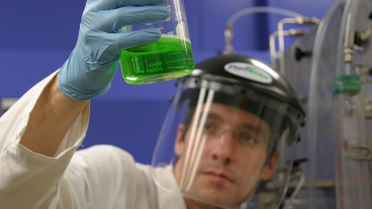 A researcher wears protective face gear and observes a beaker full of a transparent green liquid