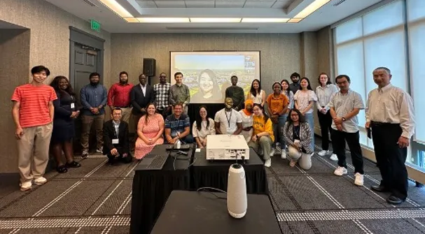 A group of people posing for a photo in a conference room with a large presentation screen displaying a virtual participant.