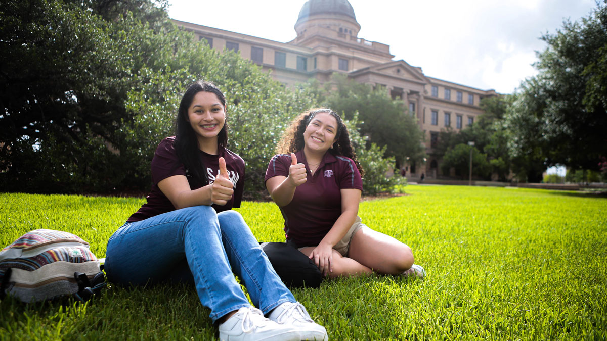 Two female students on the lawn in front of the academic building giving a "gig 'em" jesture