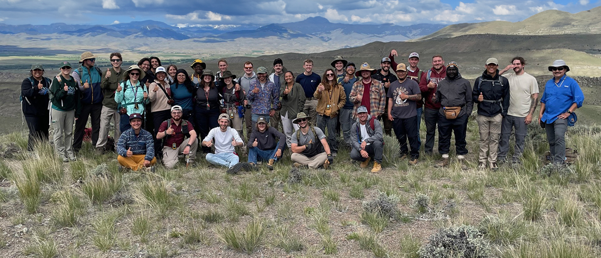 Faculty and students in a field with a valley, hills and snow capped mountains in the background