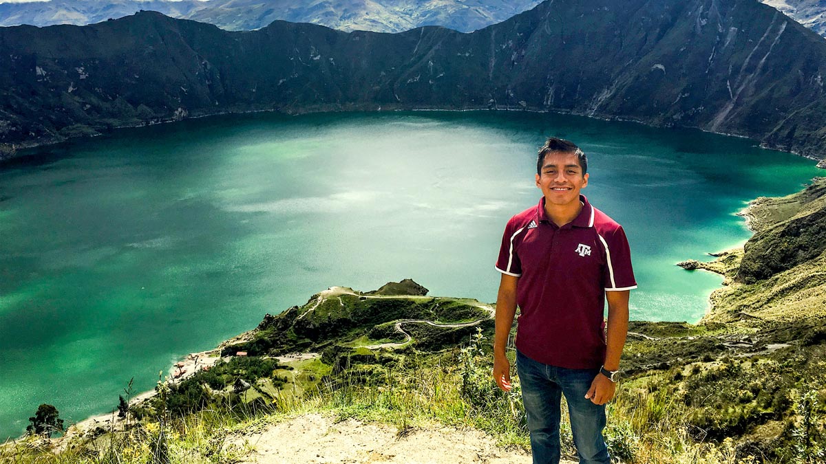 Male student posing in front of an aqua-colored body of water surrounded by mountains