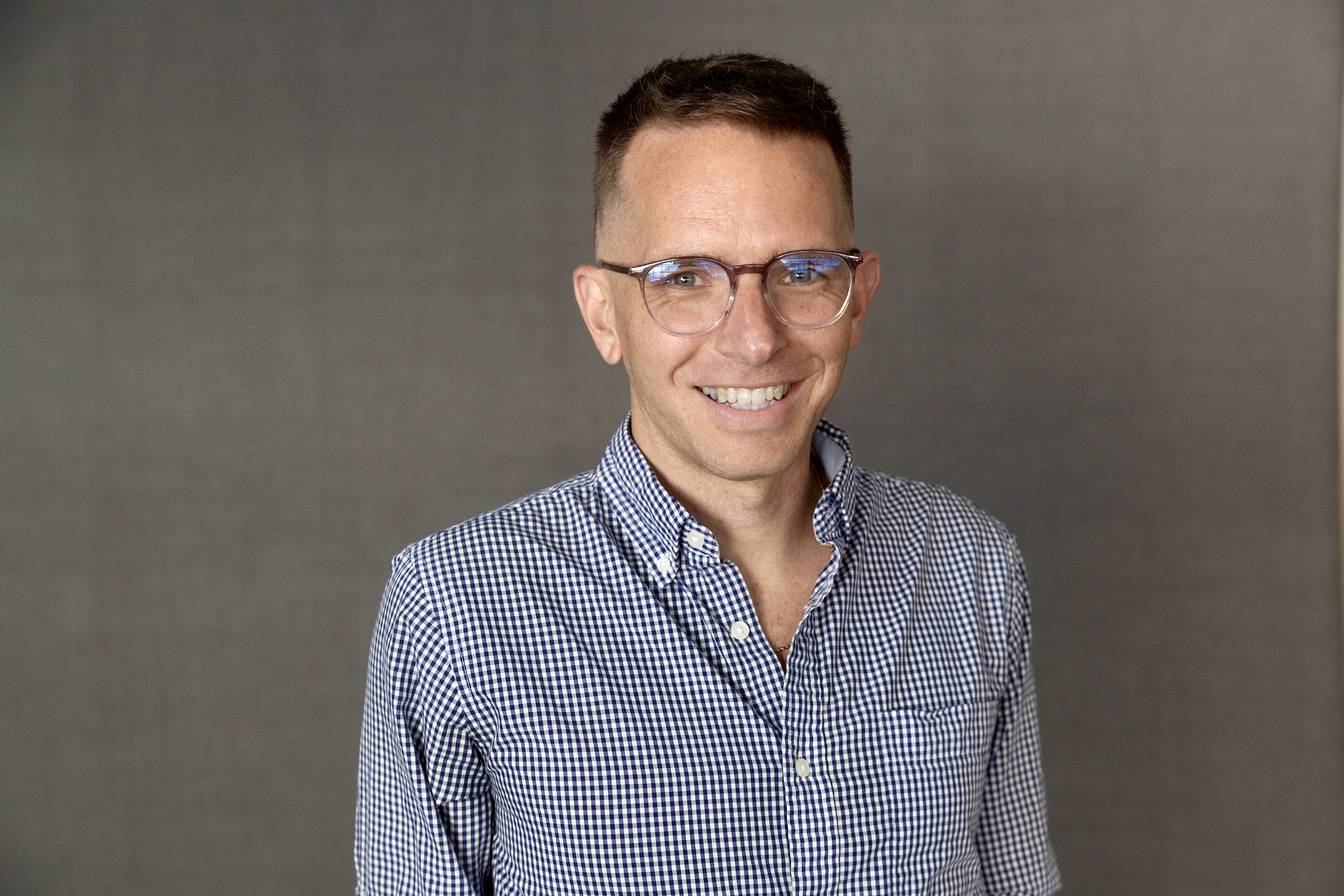 Picture of Dr. Daniel Runnels, a man with short medium brown hair, glasses, and a blue plaid button down shirt against a dark grey background.