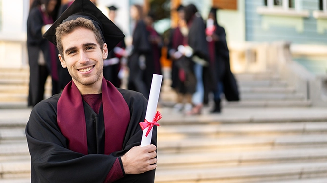 Male Graduate holding a diploma outdoors with other students in the background