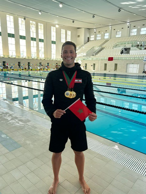 Munzy Kabbara standing in front of indoor swimming pool with four gold medals won in the Tangiers International Open swim meet.
