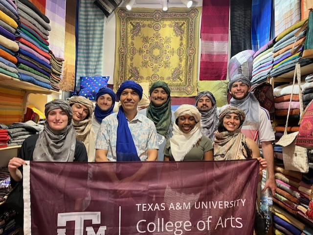 Professor Ayari with a group of Texas A&M University students wearing traditional attire and holding a college banner in a vibrant textile shop.