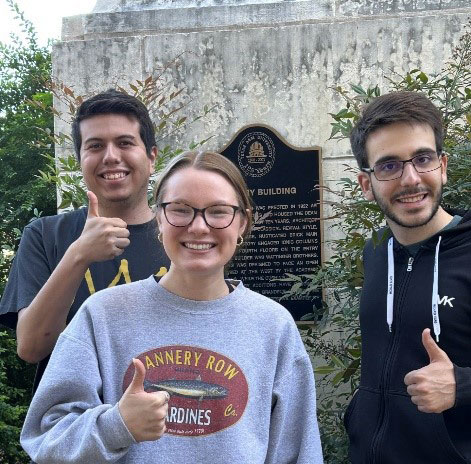 Two male students and one female student smiling and standing in front of a historical marker outside. 