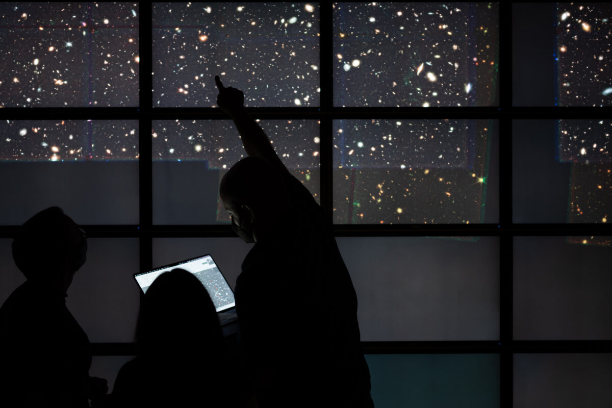 Three researchers with the Cosmic Evolution Early Release Science Survey, one of whom is holding a laptop computer and pointing above his head, stand silhouetted against a backdrop of deep field images from the James Webb Space Telescope projected onto a wall of video screens at the Texas Advanced Computing Center on The University of Texas at Austin campus