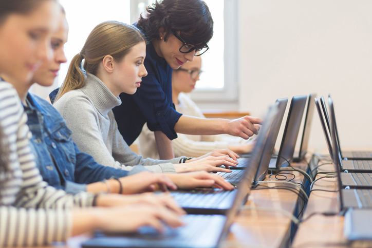 Stock image of female students working on laptops in a computer lab with teacher pointing at the computer screen