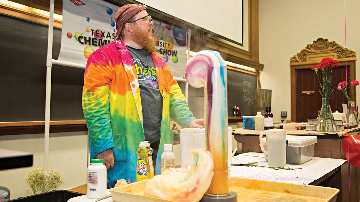 Lecture in with tie-dye lab coat and goggles on in front of lab experiment