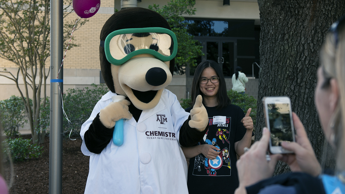 Female student posing next to mole mascot in laboratory outfit giving thumbs up with woman taking a photo of them on her cell phone