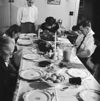 Black and white photo of a family sitting down with bowed heads to a Thanksgiving dinner with all the place settings, centerpiece, tablecloth and turkey with side dishes