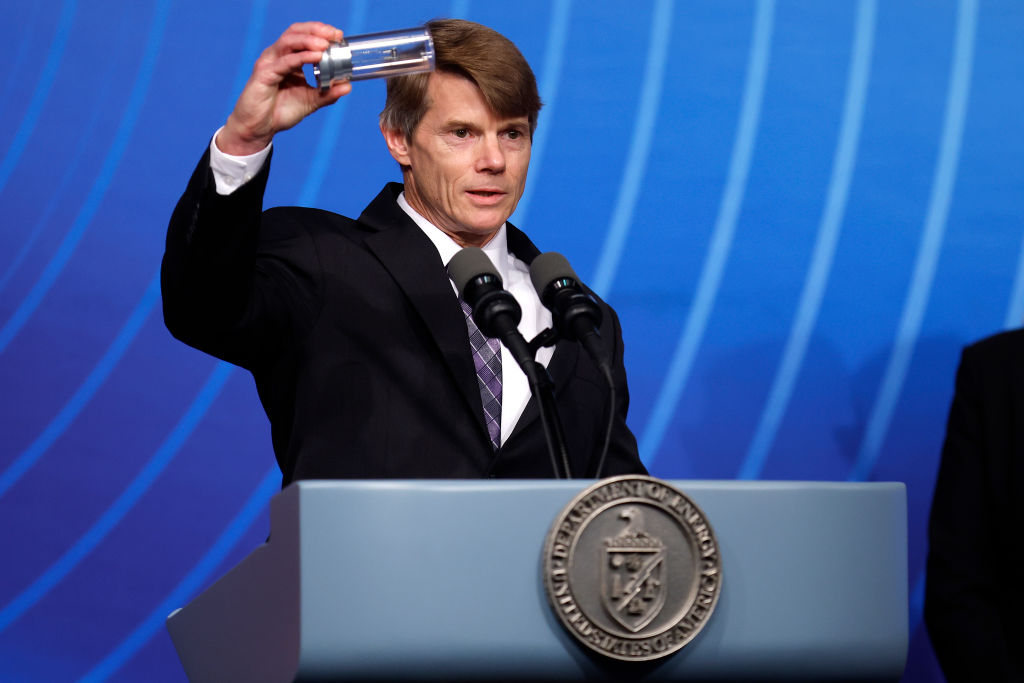 National Nuclear Security Administration Deputy Administrator for Defense Programs Marvin Adams holds up a cylinder while standing at a podium during a Department of Energy news conference