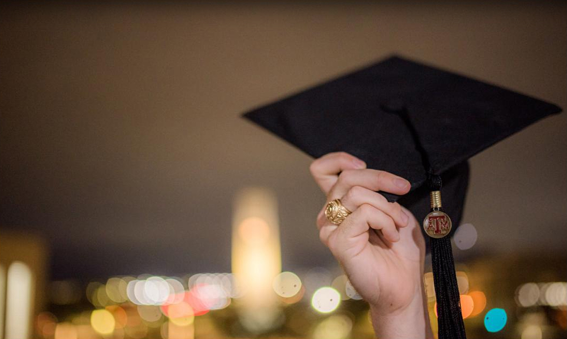 Close-up of the hand of a Texas A&amp;M graduate wearing an Aggie Ring and holding up his graduation cap against a nighttime skyline featuring Albritton Tower in the background