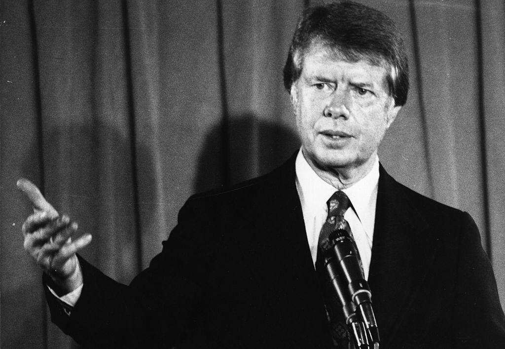 Former U.S. President Jimmy Carter speaks at a podium with his right arm and hand outstretched in illustration of a point