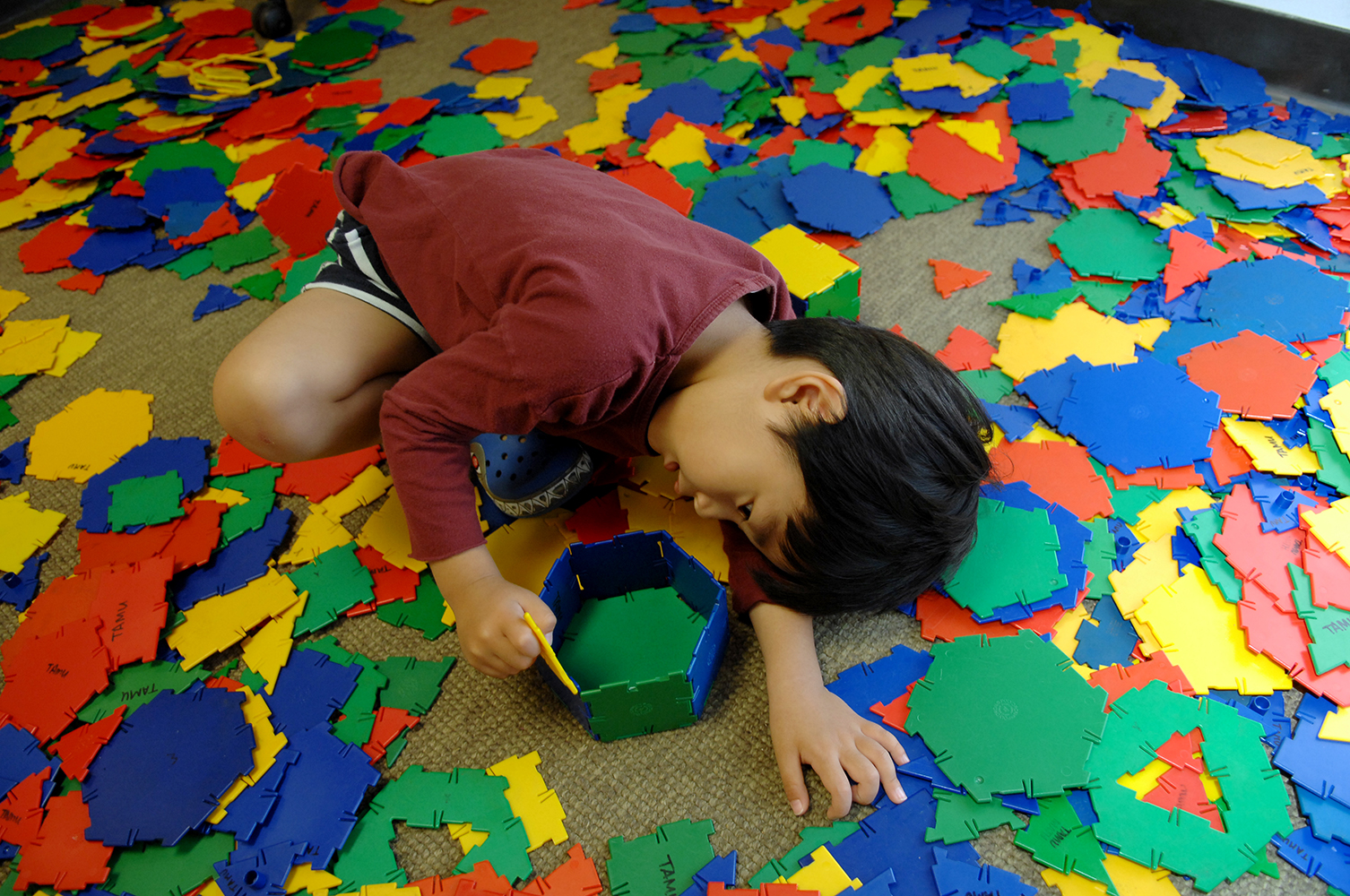 An elementary student sits and builds a tower on the floor with colorful tiles strewn around him