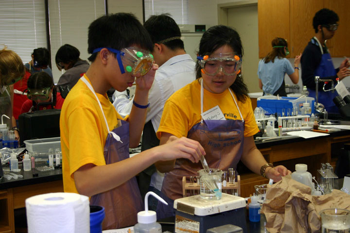 Two students wear safety goggles and aprons and heat liquid on a Bunsen burner