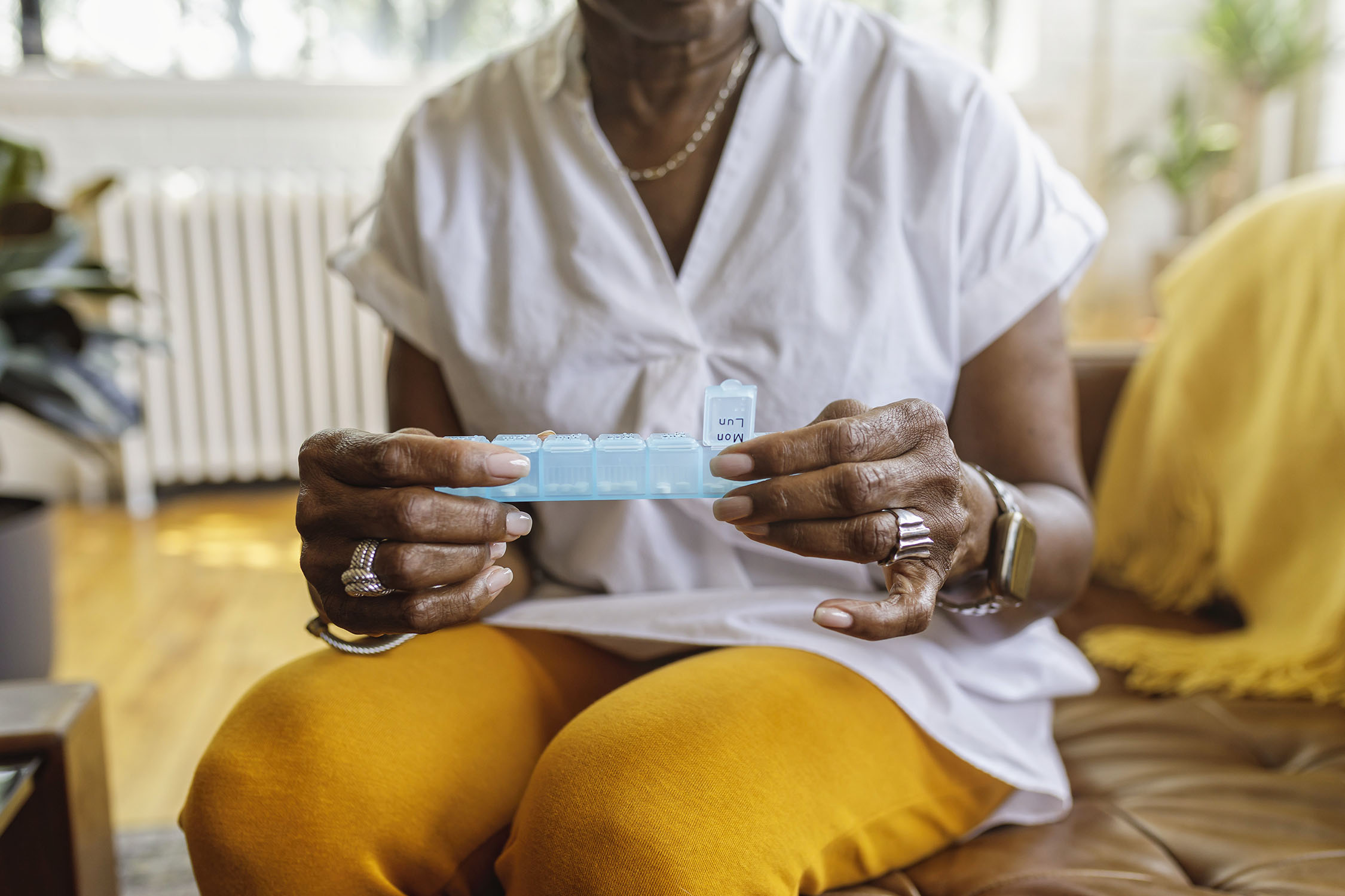 A senior black woman sits on the couch at home and takes medications from a daily pill organizer