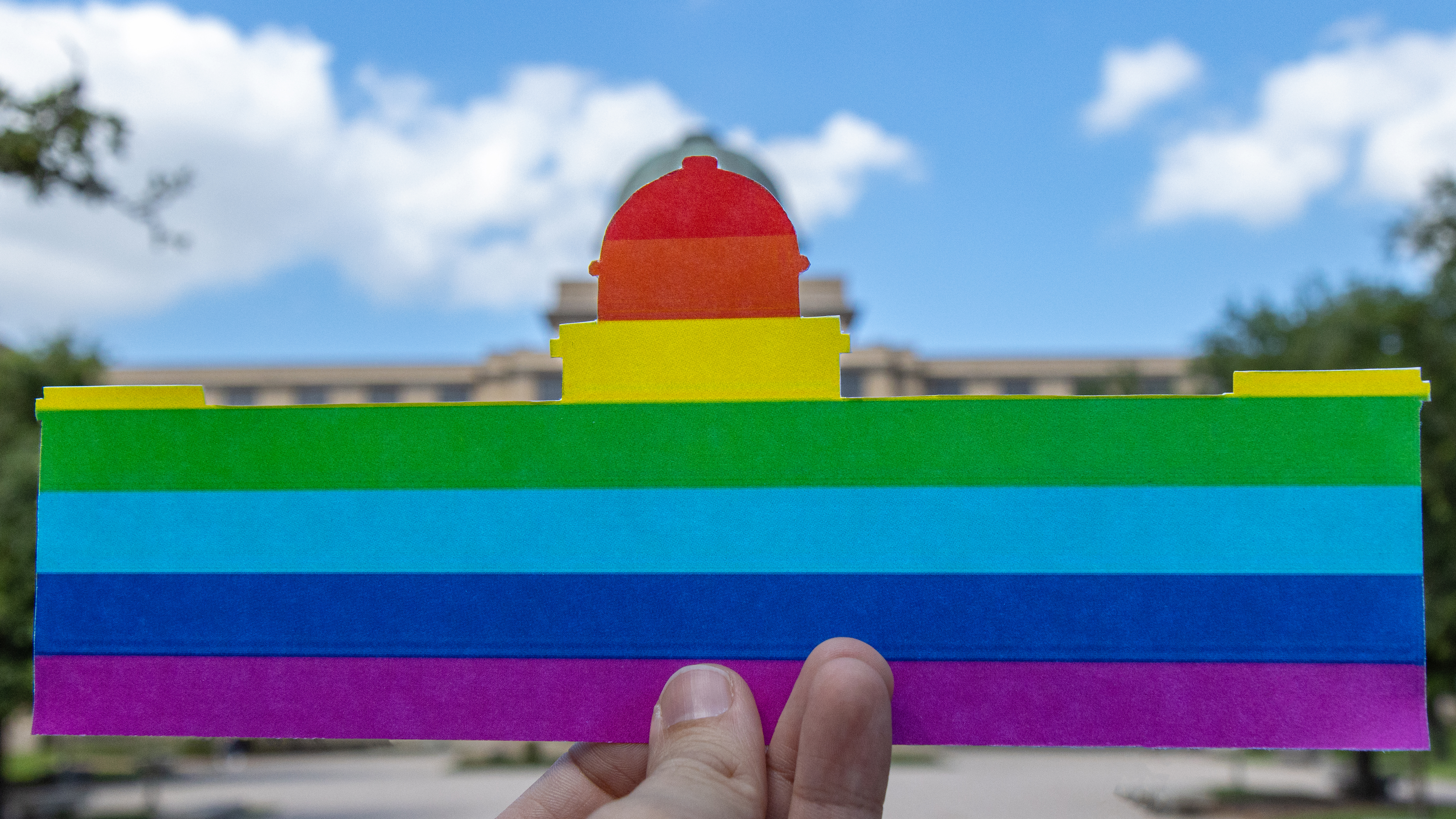 Hand holds up Pride Flag-printed cutout of Academic Building over the actual Academic Building. 