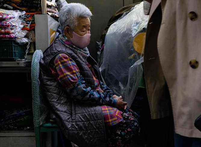 An elderly woman wearing a mask sits in a chair as people walk by her in the marketplace