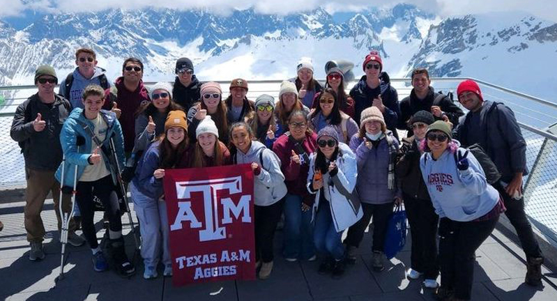 Texas A&amp;M students participating in the 2023 Student Experiences Abroad in Meteorology program pose with a maroon Texas A&amp;M University flag with the Alps in the background