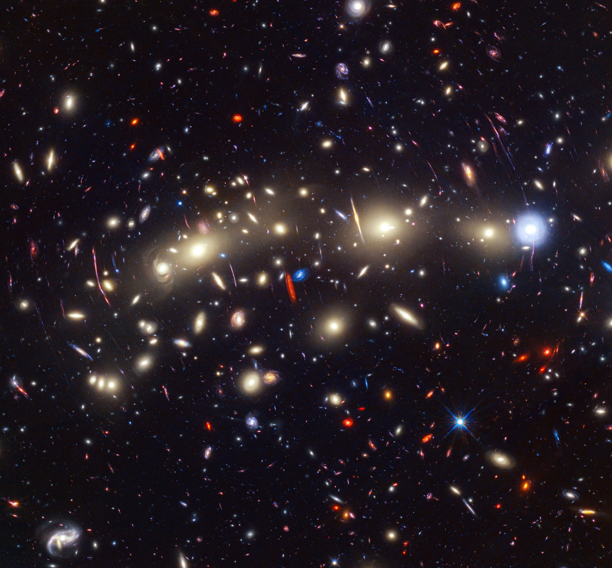 Panchromatic view of galaxy cluster MACS0416 featuring a vivid landscape of galaxies and bright individual stars. The image was created by combining infrared observations from NASA’s James Webb Space Telescope with visible-light data from NASA’s Hubble Space Telescope, resulting in a prismatic panorama of blues and reds – colors that give clues to the distances of the galaxies.