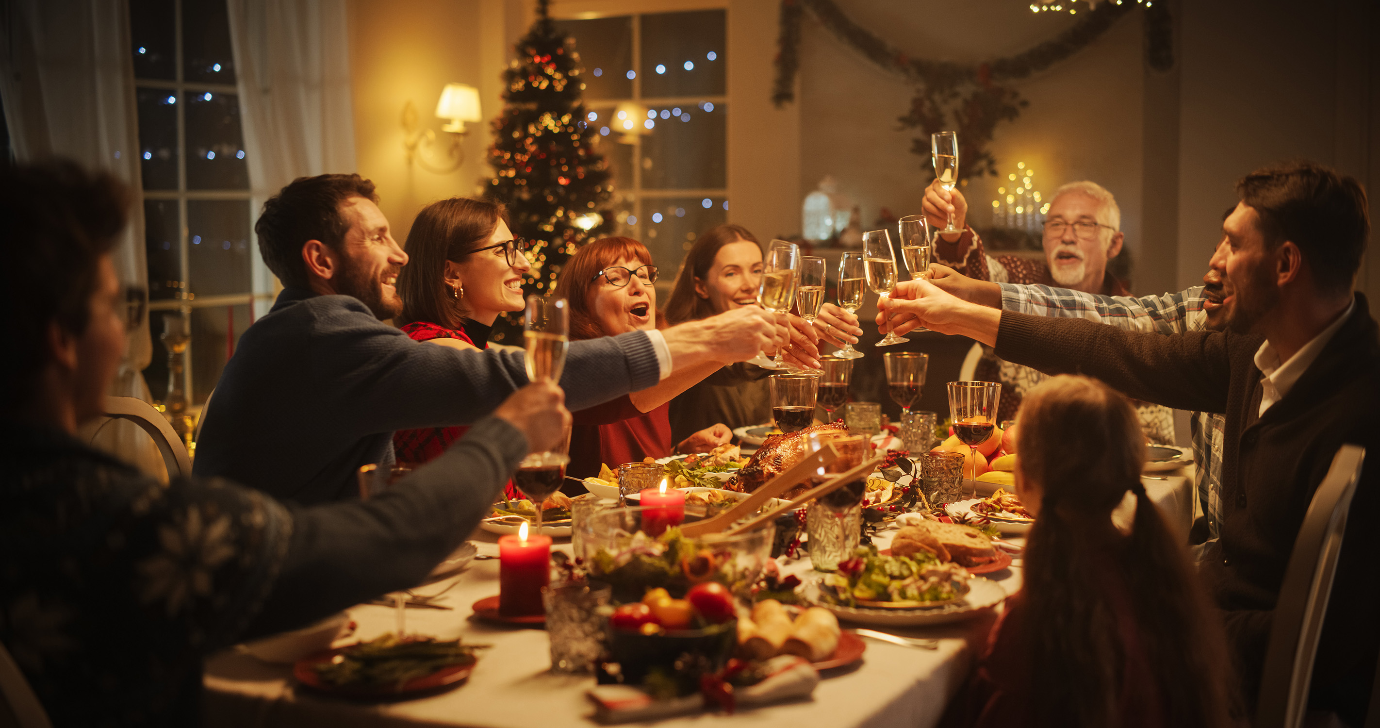 A mixed group of people seated around a rectangular table in preparation to enjoy the holiday meal set before them clink raised wine glasses in a celebratory toast in a room decked with holiday decor