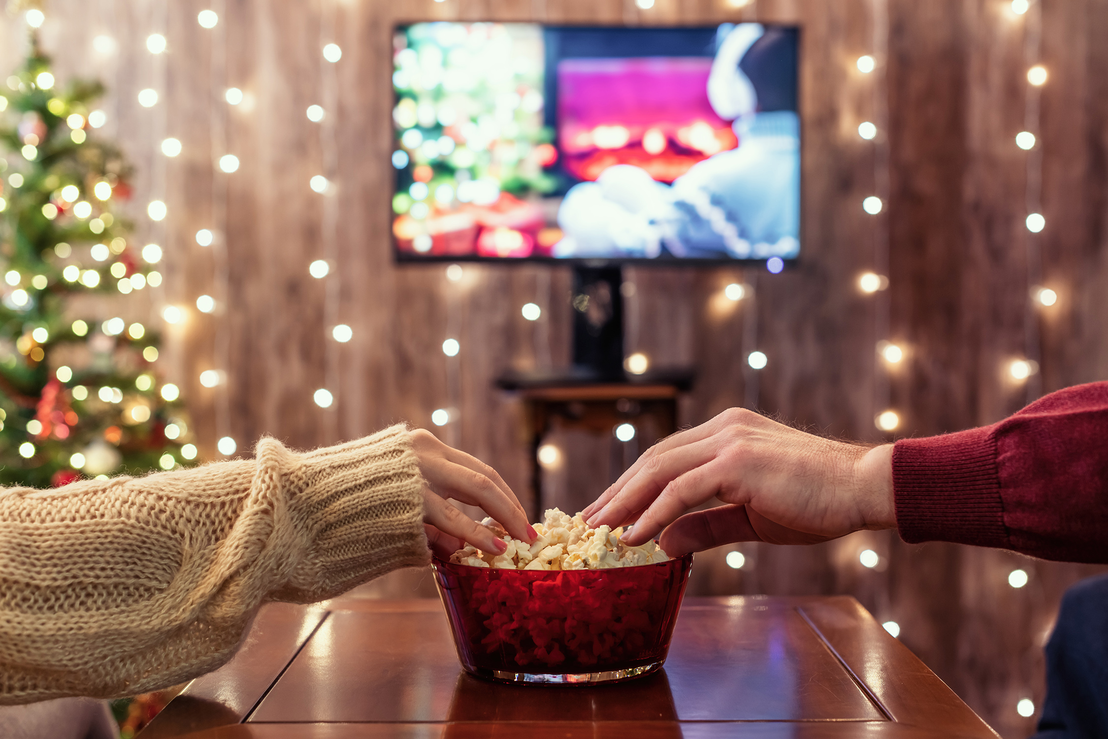Two people reach for a bowl of popcorn while sitting on opposite sides of a small wooden table and watching television
