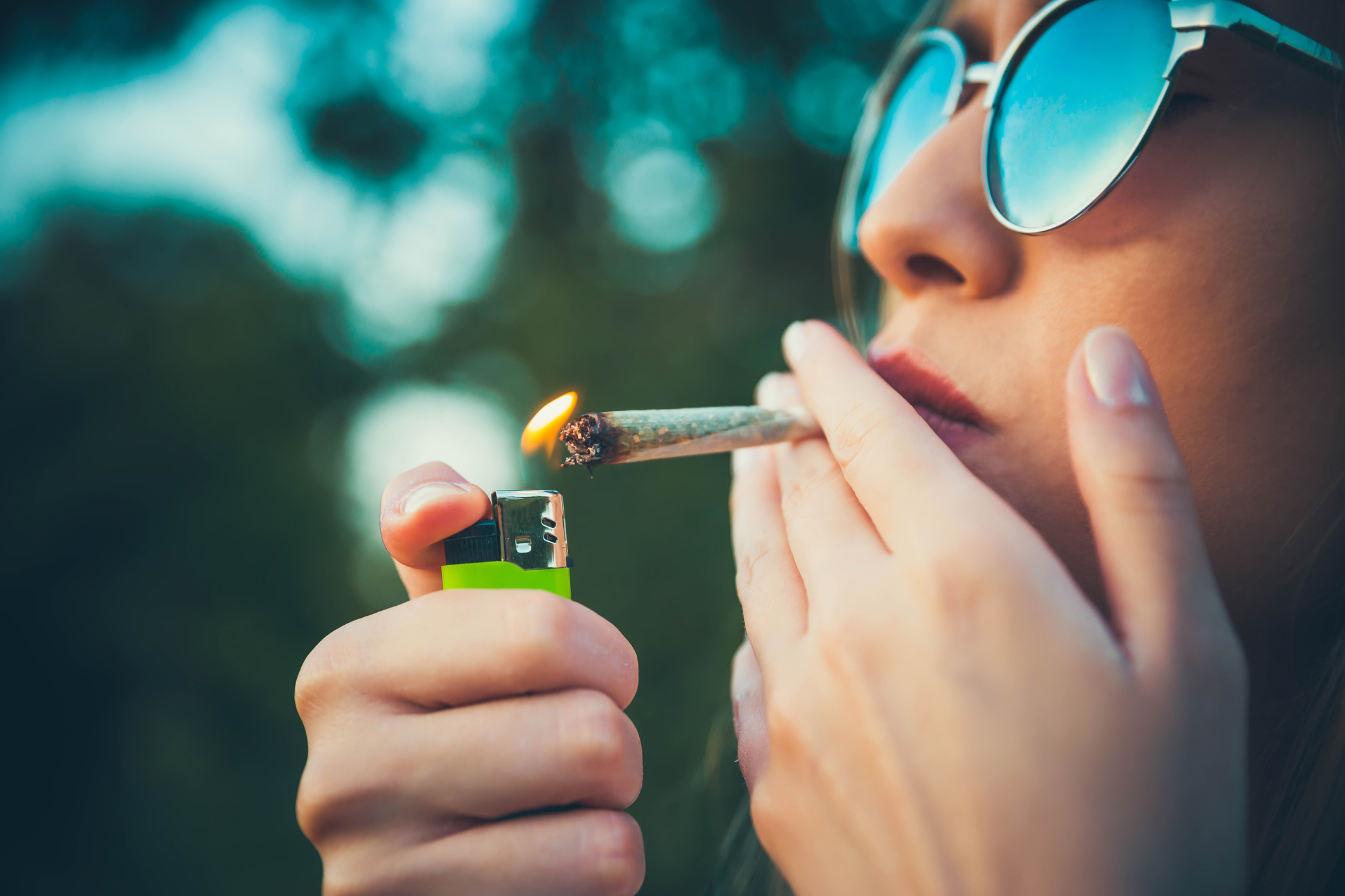 Young woman wearing sunglasses shown in profile lighting up a marijuana cigarette 