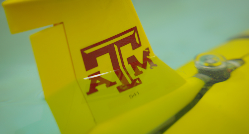 A yellow glider featuring a maroon Texas A&M University logo floats in a pool of water within a Geochemical and Environmental Research Group laboratory
