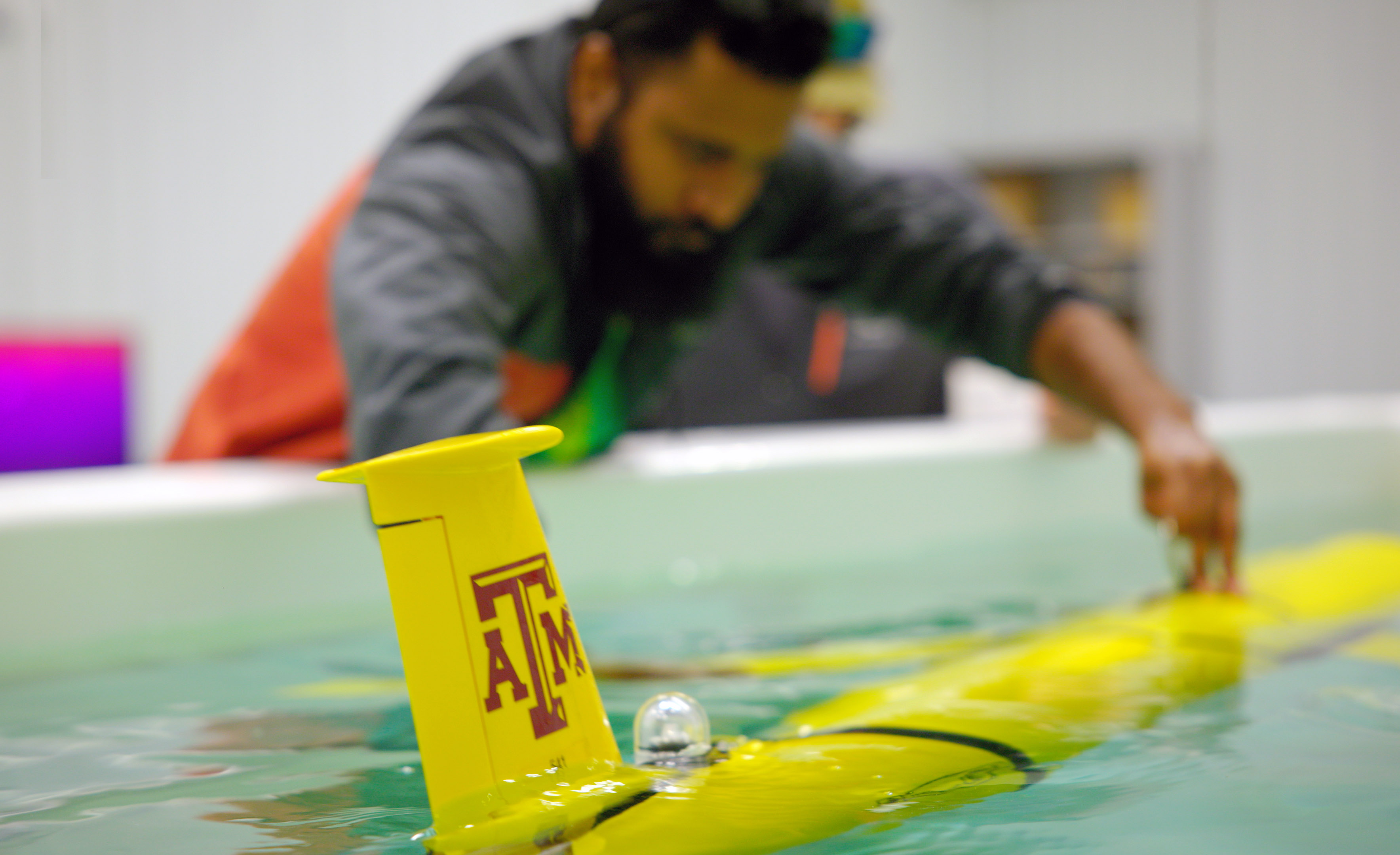 Postdoctoral researcher Sakib Mahmud  observes a yellow glider featuring a maroon Texas A&amp;M University logo as it floats in a pool of water within a Geochemical and Environmental Research Group laboratory