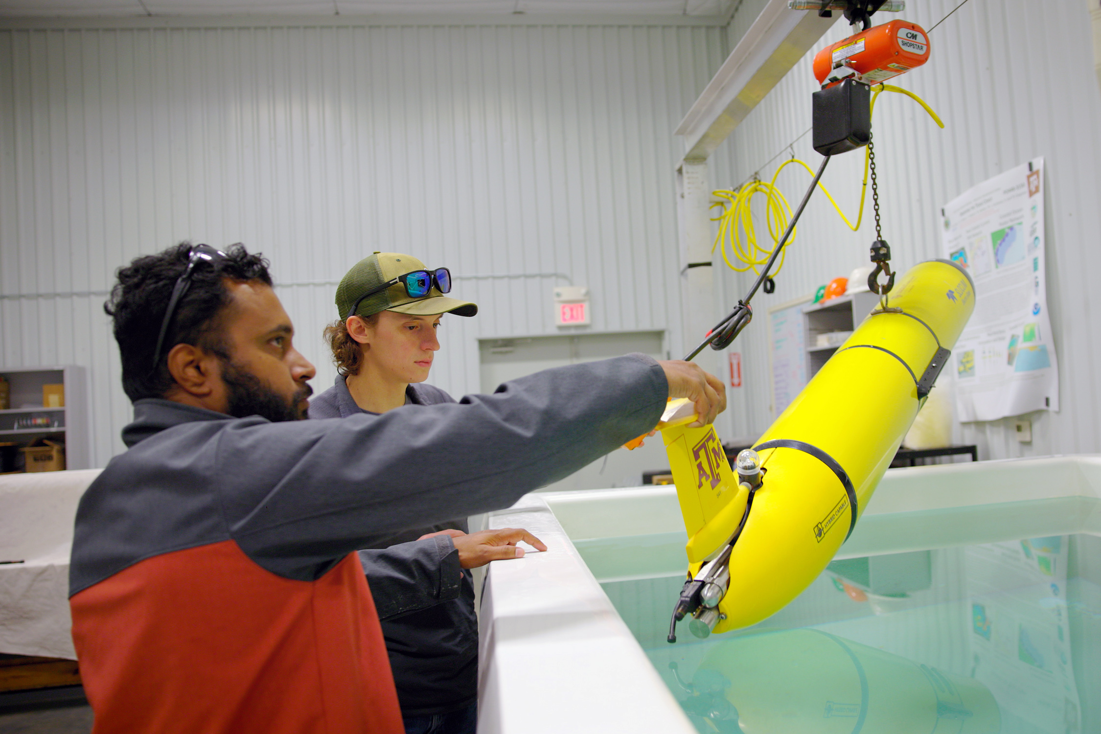 Two Geochemical and Environmental Research Group members observe a yellow glider featuring a maroon Texas A&amp;M University logo as it floats in a pool within the GERG laboratory
