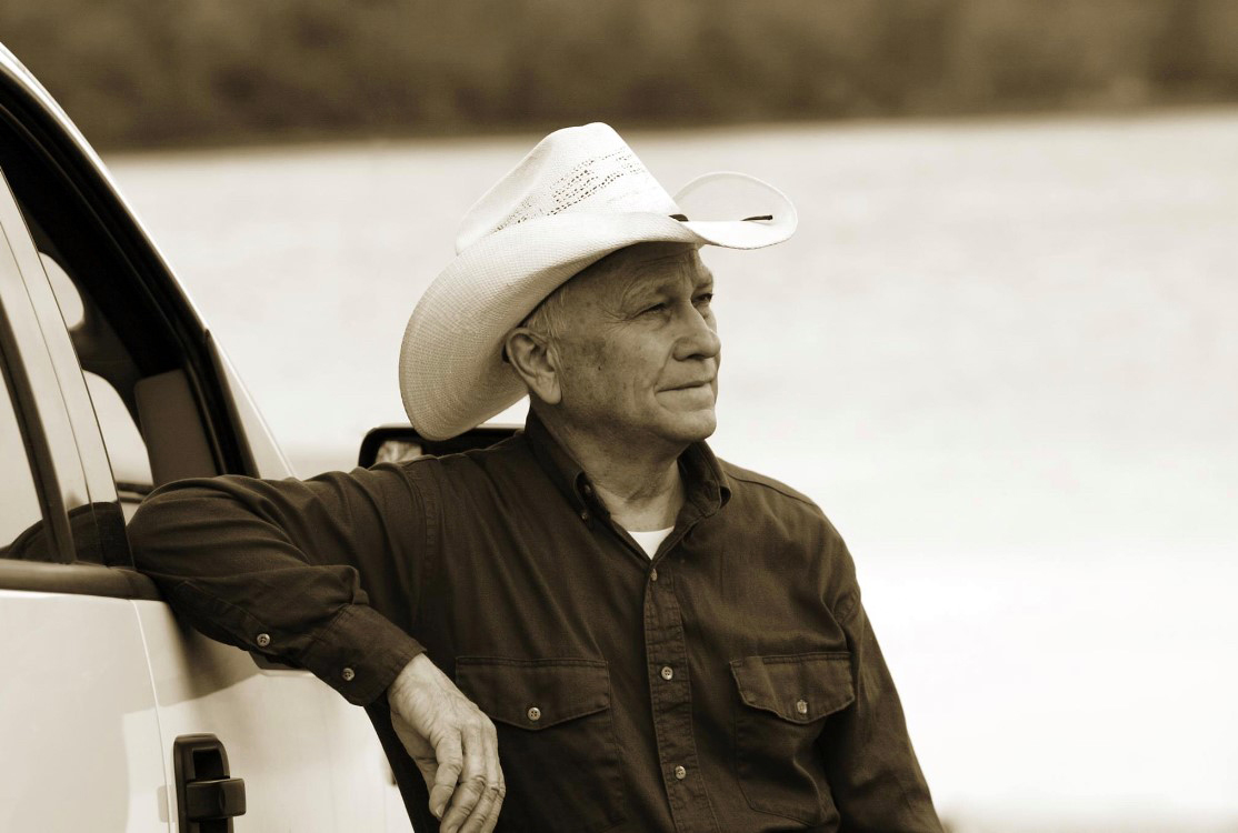 Texas A&M University economics professor Thomas Saving, wearing a straw hat and staring off into the horizon while leaning an elbow on the open window frame of his pickup
