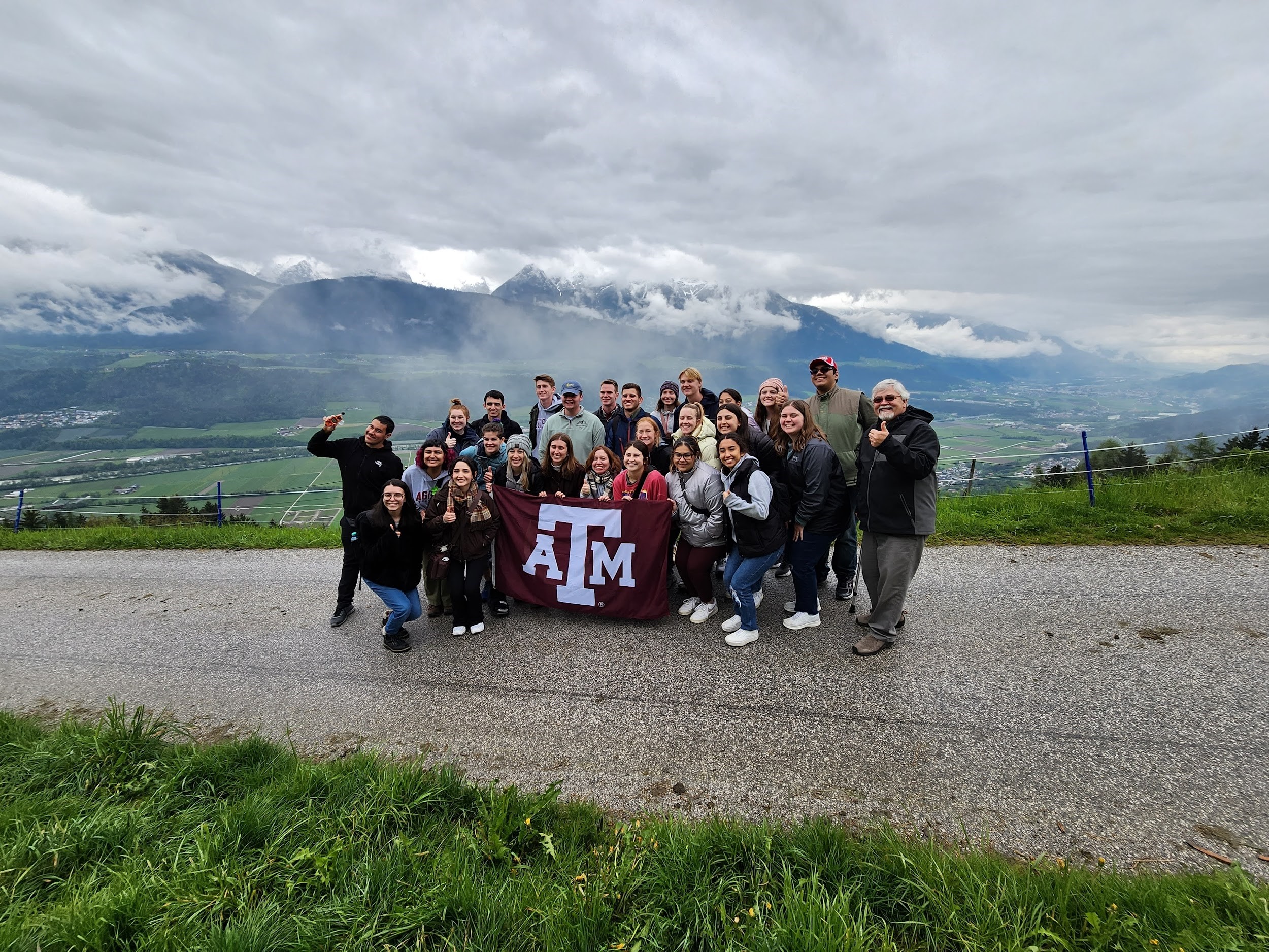 Texas A&amp;M University students participating in the 2023 Student Experiences Abroad in Meteorology program pose with a maroon Texas A&amp;M University flag with the Alps in the background