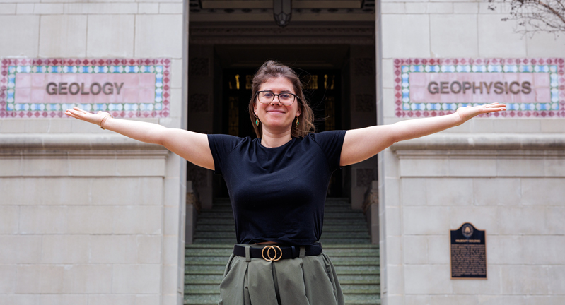 Texas A&M University Geology and Geophysics professor Brandi Lenz stands outside the Halbouty Building with outstretched arms, extending her palms in metaphorical support of the words "Geology" and "Geophysics" that appear in intricate tile format on the building's exterior wall