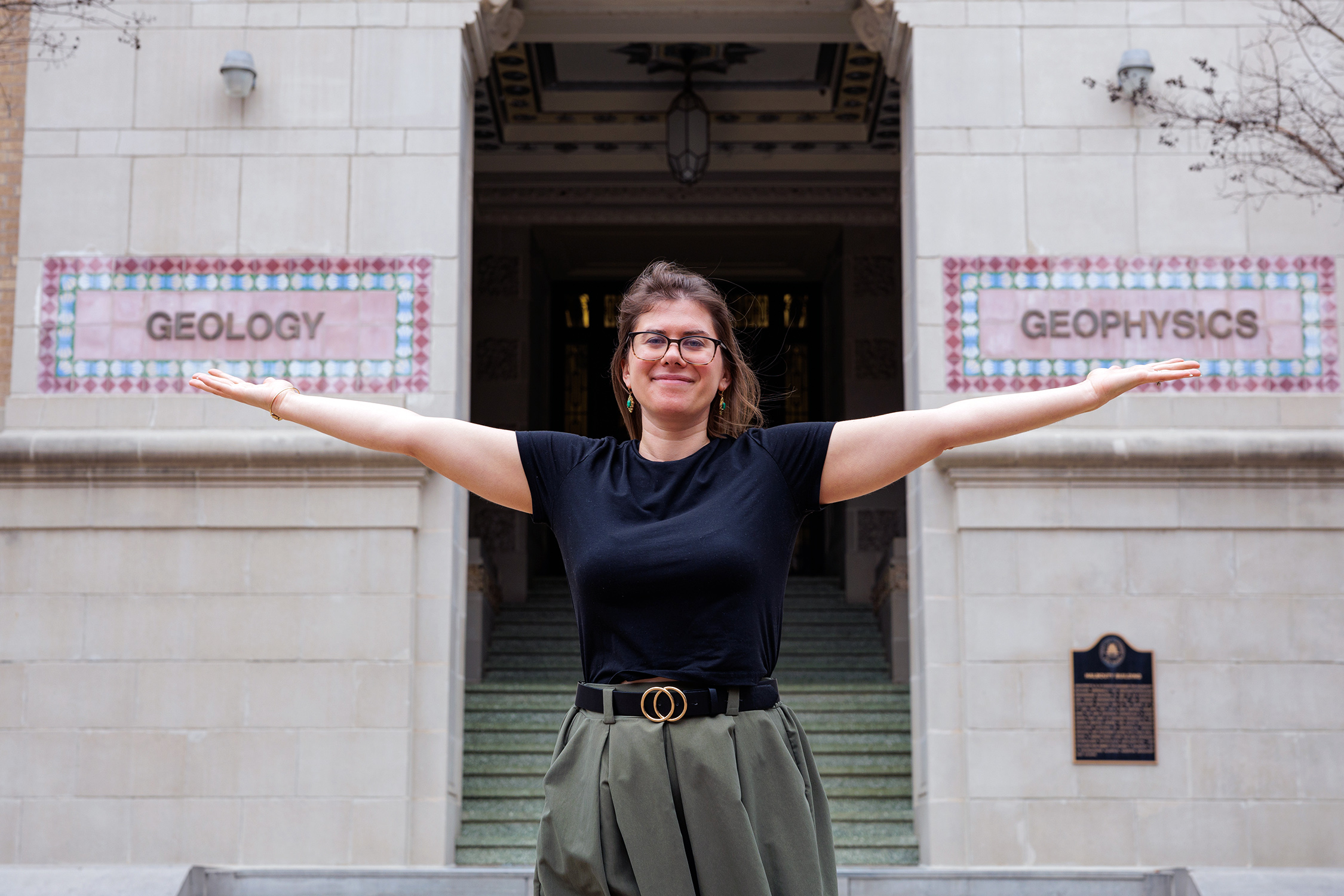 Texas A&amp;M University Geology and Geophysics professor Brandi Lenz stands outside the Halbouty Building with outstretched arms, extending her palms in metaphorical support of the words "Geology" and "Geophysics" that appear in intricate tile format on the building's exterior wall