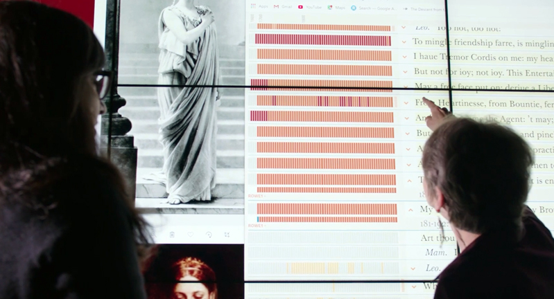 Texas A&M University English Professor Laura Mandell extends her index finger toward a wall-sized touch-screen display featuring the New Variorum Shakespeare website in the Humanities Visualization Space within the Center of Digital Humanities Research