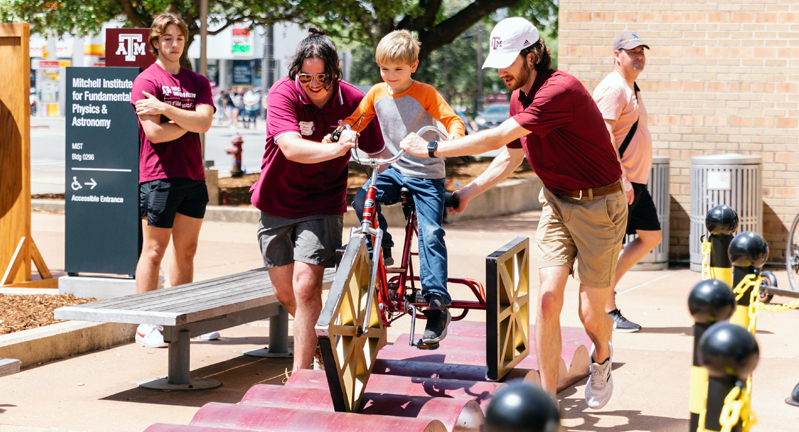 Two Texas A&M University student volunteers assist a male youth in riding a square-wheeled bicycle at the 2023 Physics and Engineering Festival, held April 1, 2023