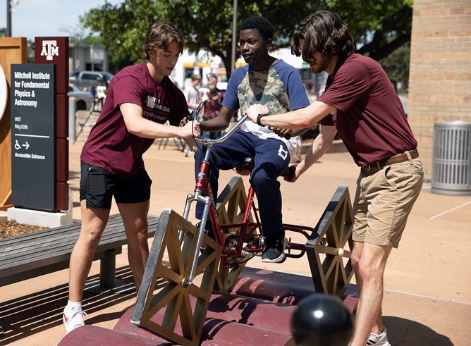 Two Texas A&amp;M University student volunteers assist a male youth in riding a square-wheeled bicycle at the 2023 Physics and Engineering Festival, held April 1, 2023