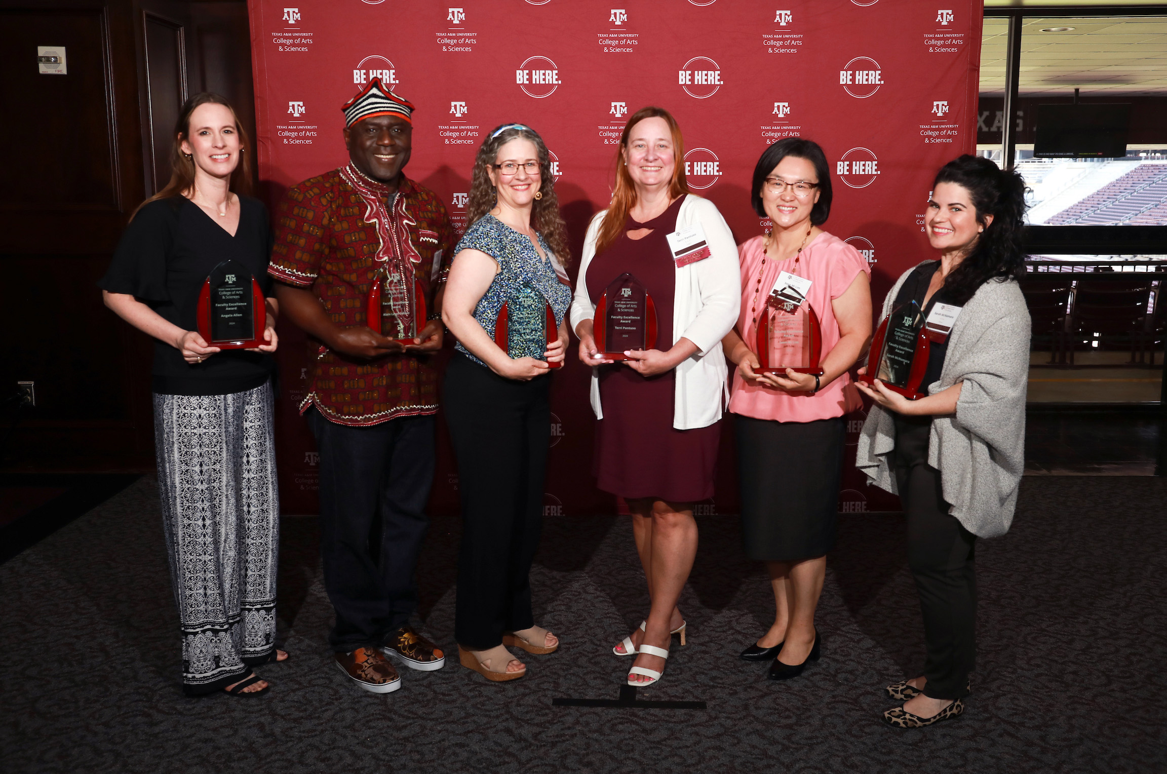 Group photograph of the six recipients of 2024 College of Arts and Sciences Faculty Excellence Awards, posing with their plaques in front of a maroon backdrop featuring the college logo and the "Be Here" stamp in white