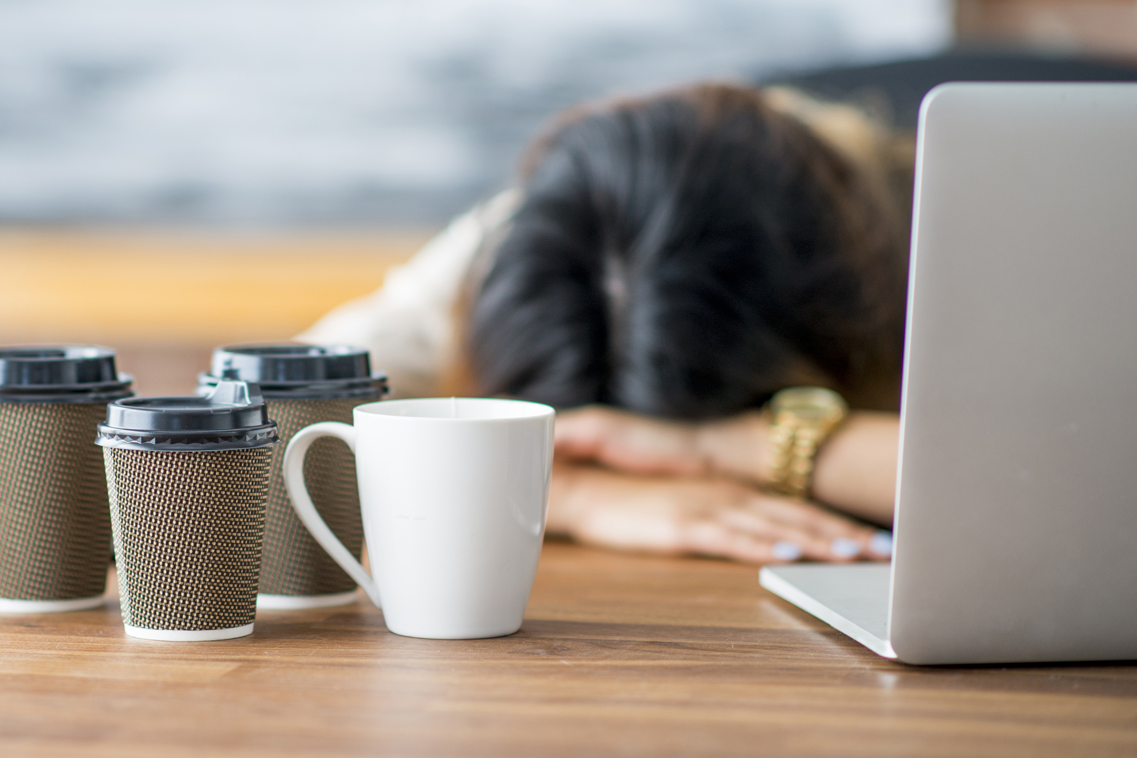 Exhausted female sleeps with her head on her desk, flanked by a laptop and multiple disposable coffee cups