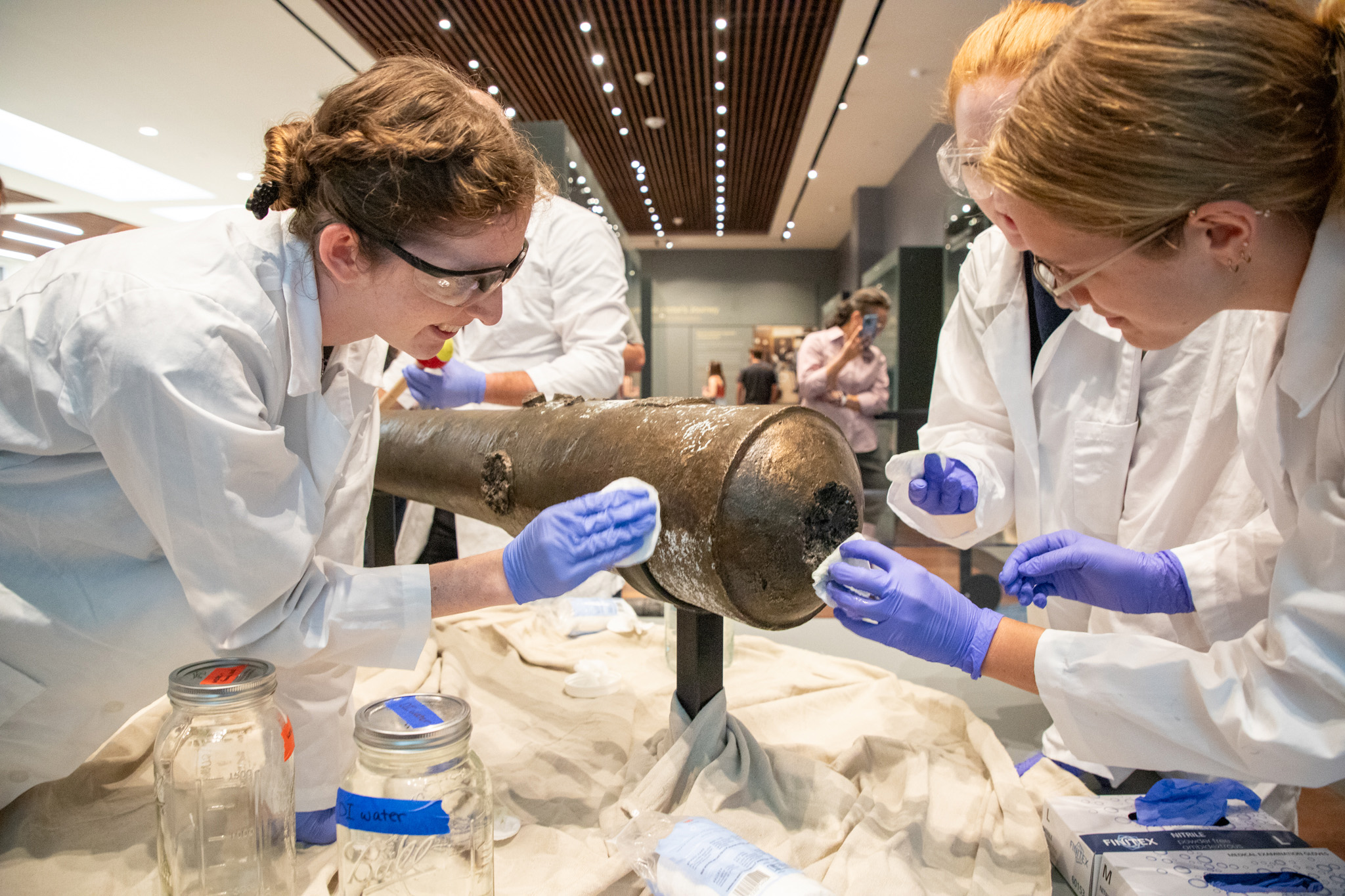 Four female graduate students from Texas A&amp;M University wearing white lab coats, goggles and blue latex gloves work to clean a bronze cannon at the Alamo Museum in San Antonio