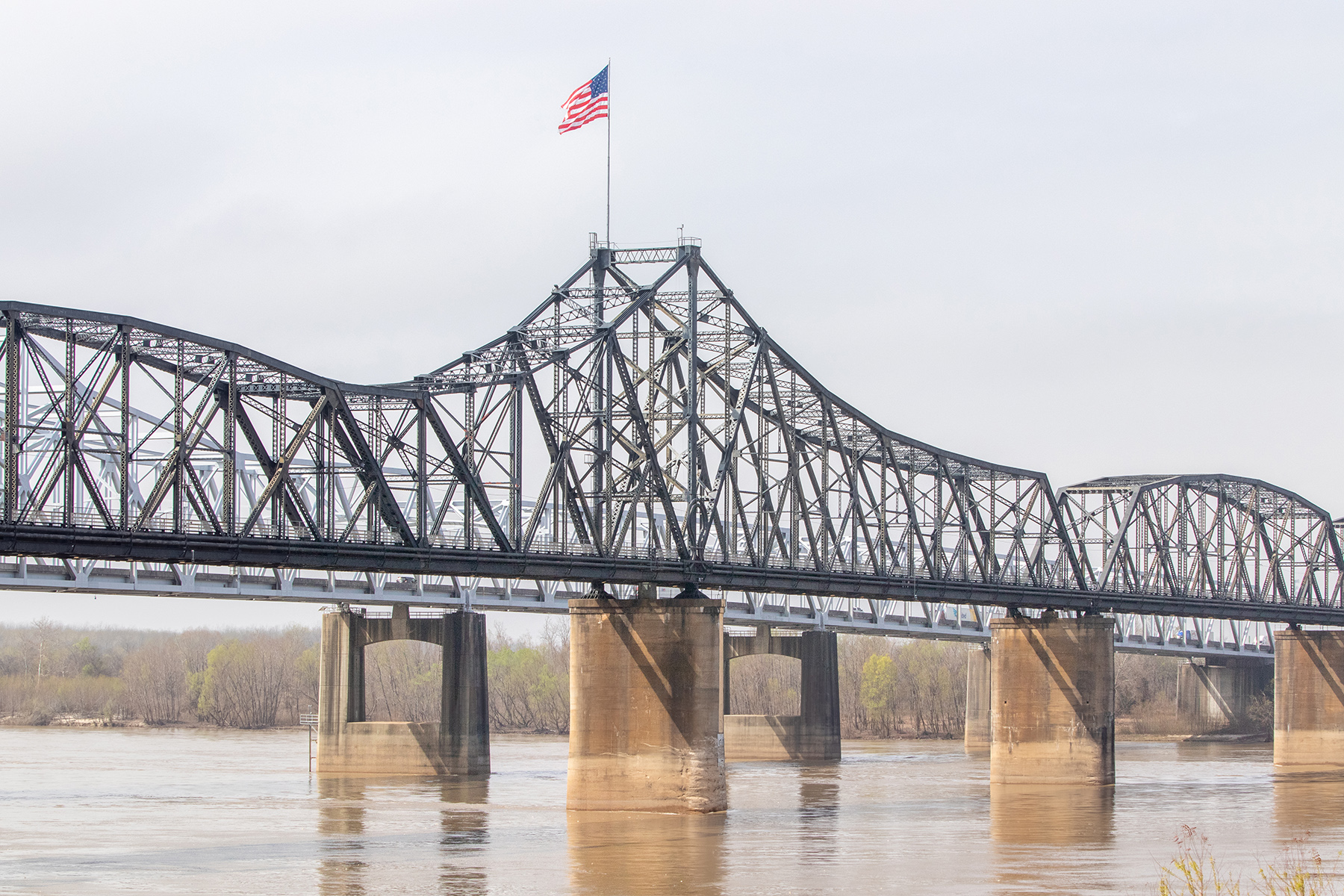 A view of the Vicksburg Bridge with Louisiana behind it. The American Flag proudly flies on top of it on a beautiful sunny day.