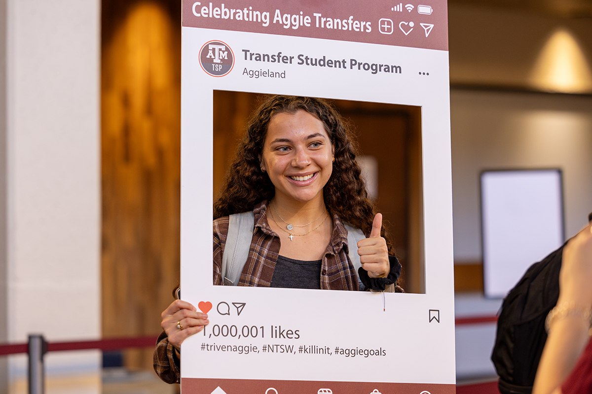 A student smiles for the camera and flashes a gig 'em thumbs up while behind a social media photo booth-style prop during an event for transfer students at Texas A&amp;M University