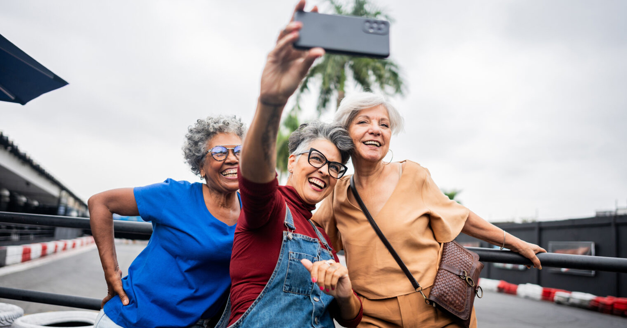 Three older females smile as the one in the center holds up a cell phone to take a selfie while standing at the outer edges of a racetrack