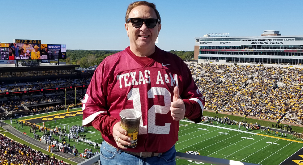 Person in a Texas A&M jersey giving a thumbs-up at a football game between Missouri Tigers and Texas A&M, with a stadium filled with spectators in the background.