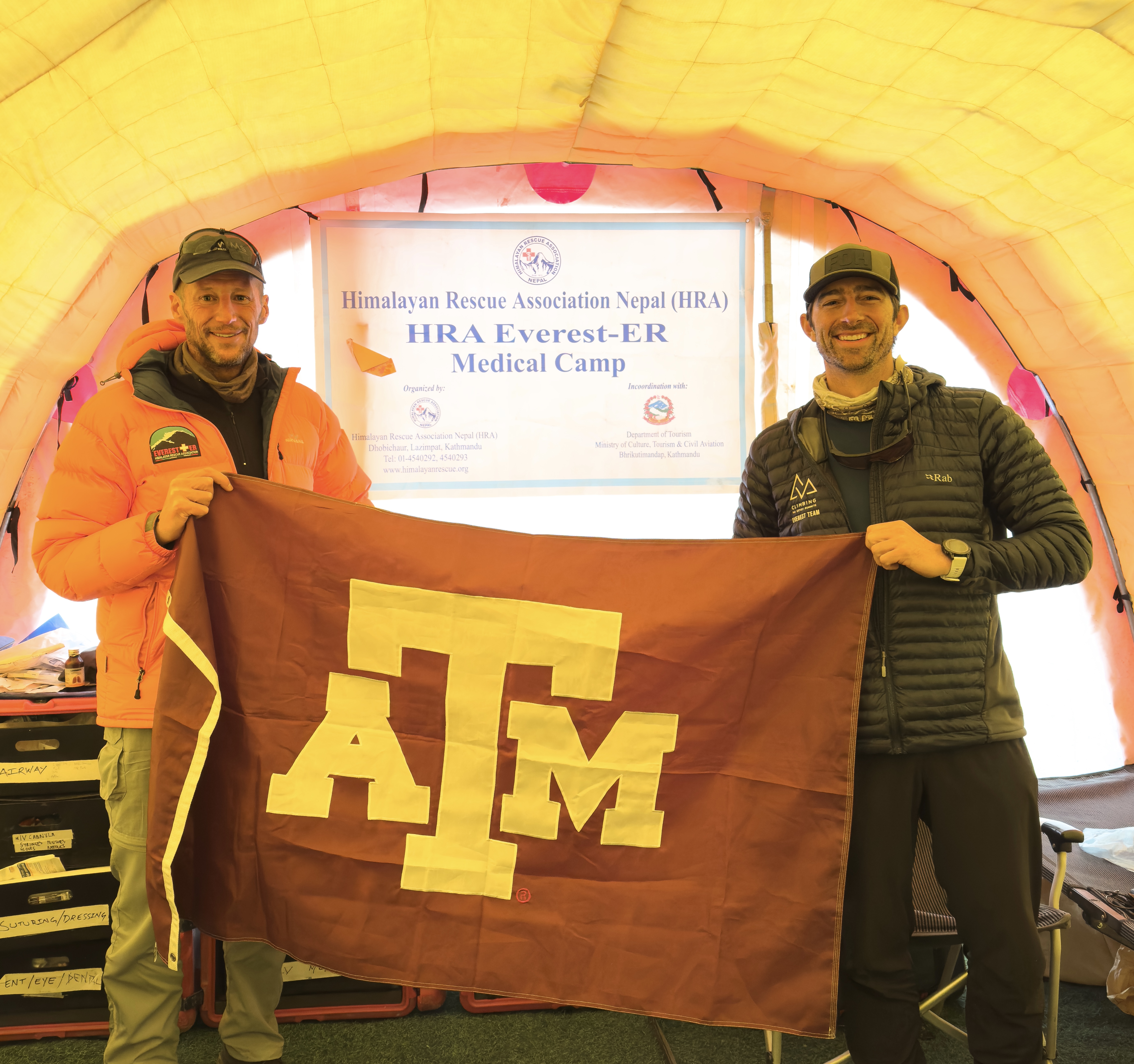 Two individuals holding a Texas A&amp;M flag initials inside a tent at the Himalayan Rescue Association Nepal (HRA) Everest-ER Medical Camp.